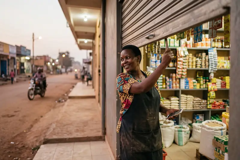 A shop owner pulling down her shutter at dusk, smiling over her shoulder with her phone in hand